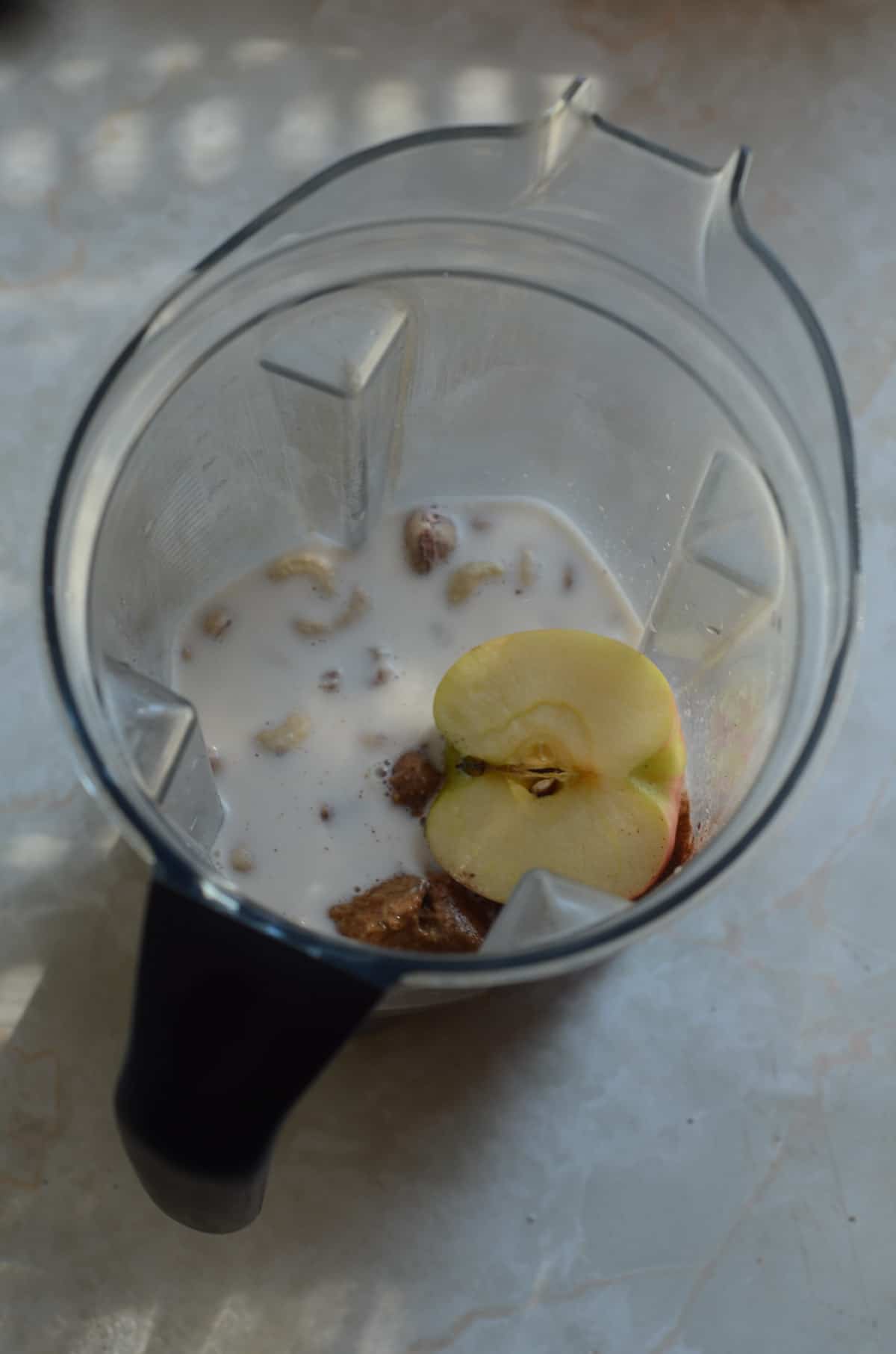 Ingredients like cashews, cinnamon, and half an apple in the blender container ready to be blended.