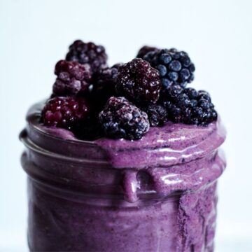 Purple smoothie in a glass jar spilling over the side with blackberries on top.