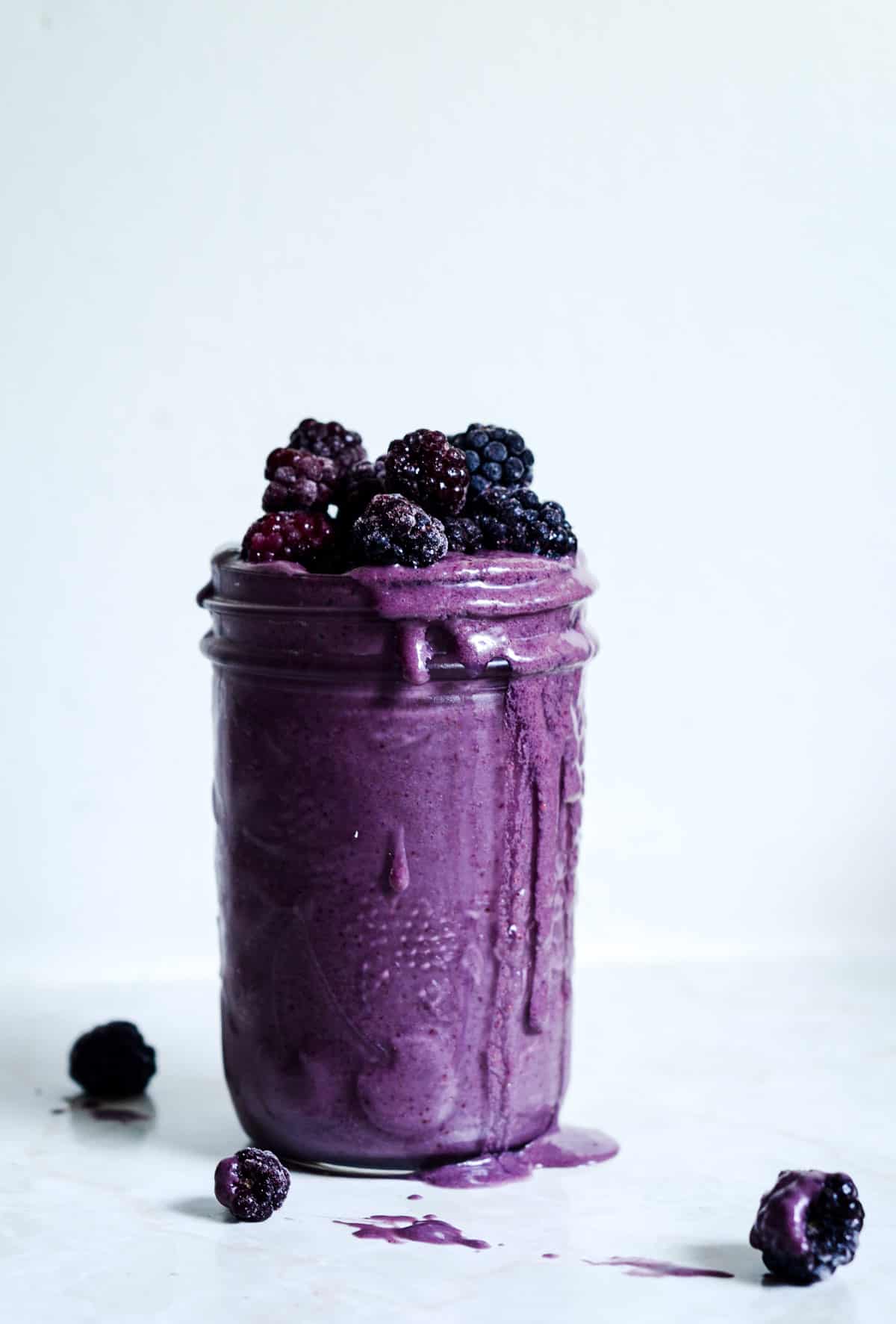 Dark purple smoothie in a glass jar with blackberries spilling out of the top.