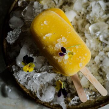 2 homemade mango popsicle on a stick with a plate of ice and pansy flowers.