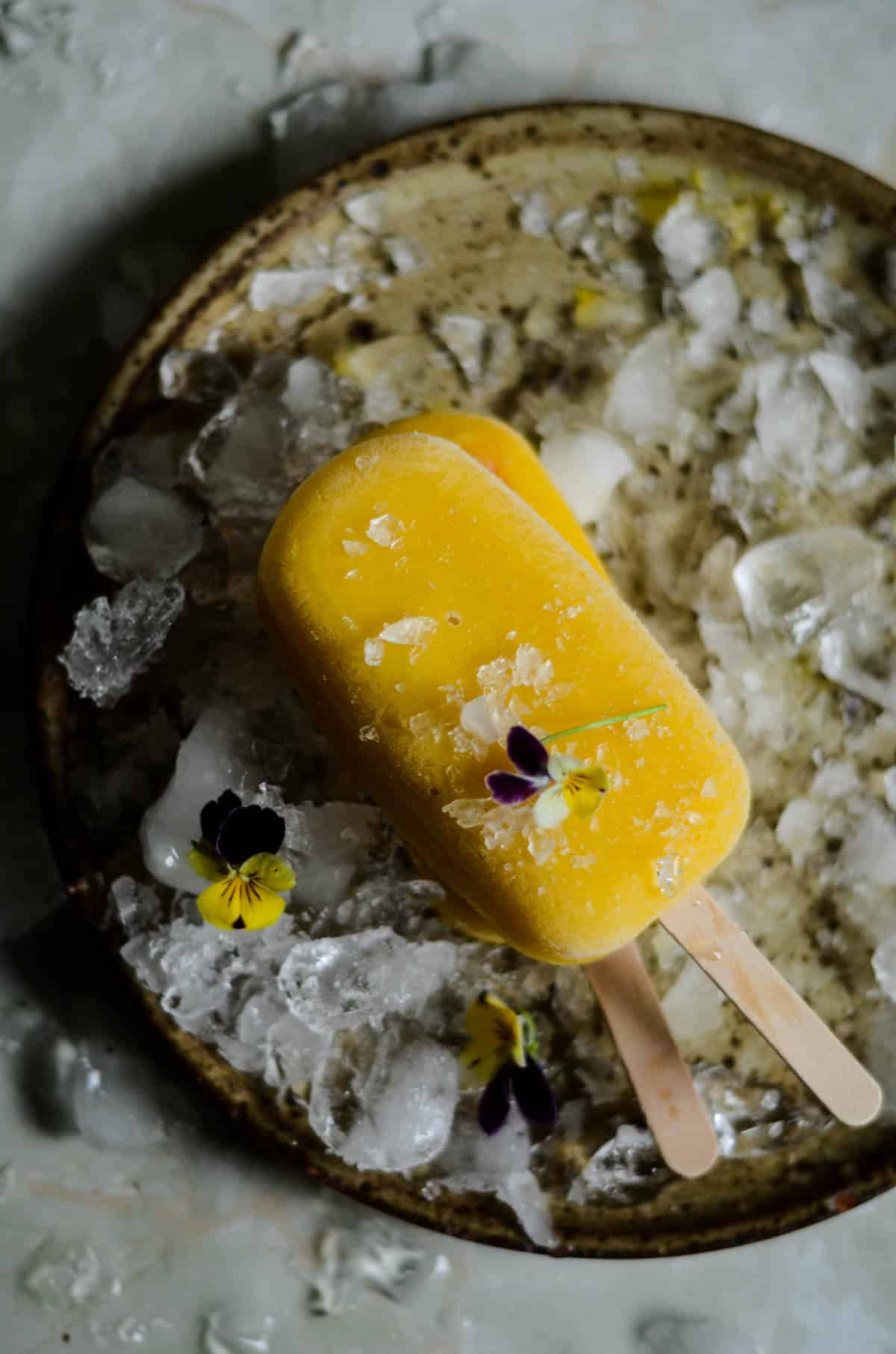 Bright yellow easy homemade mango popsicle on a stick with a plate of ice and pansy flowers.