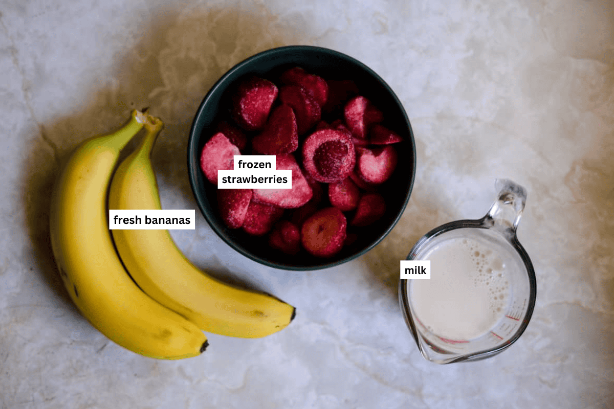 Two fresh bananas, a bowl of frozen strawberries chunks, and a measuring cup with milk.
