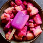Pink frozen popsicle with bite out of it sitting in a bowl of pink and yellow frozen fruit.