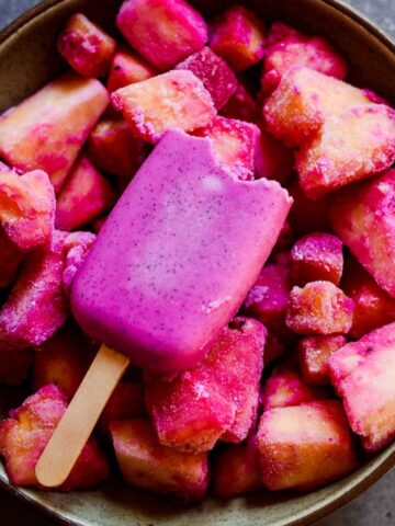 Pink frozen popsicle with bite out of it sitting in a bowl of pink and yellow frozen fruit.