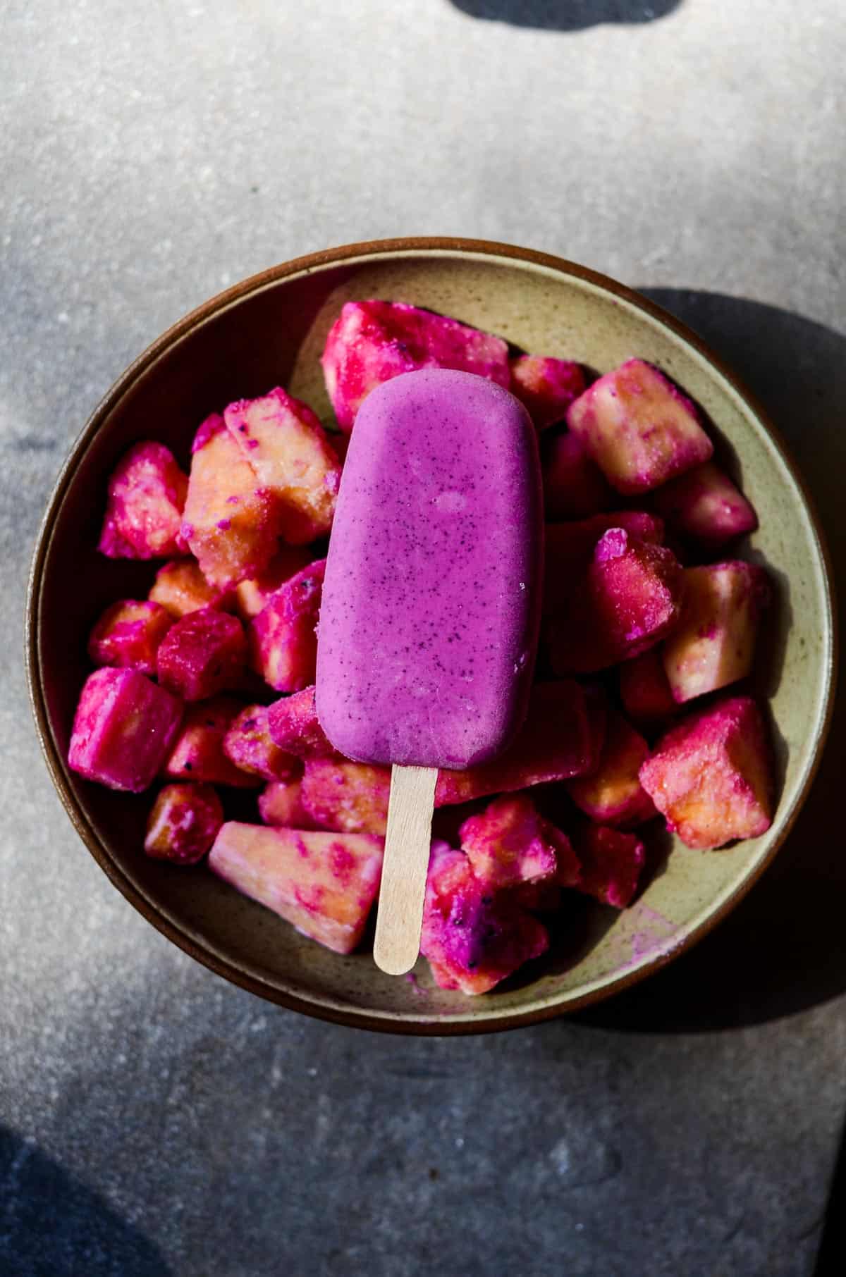   Bright purple popsicle sitting on top of frozen tropical fruit, pineapple, in a bowl.