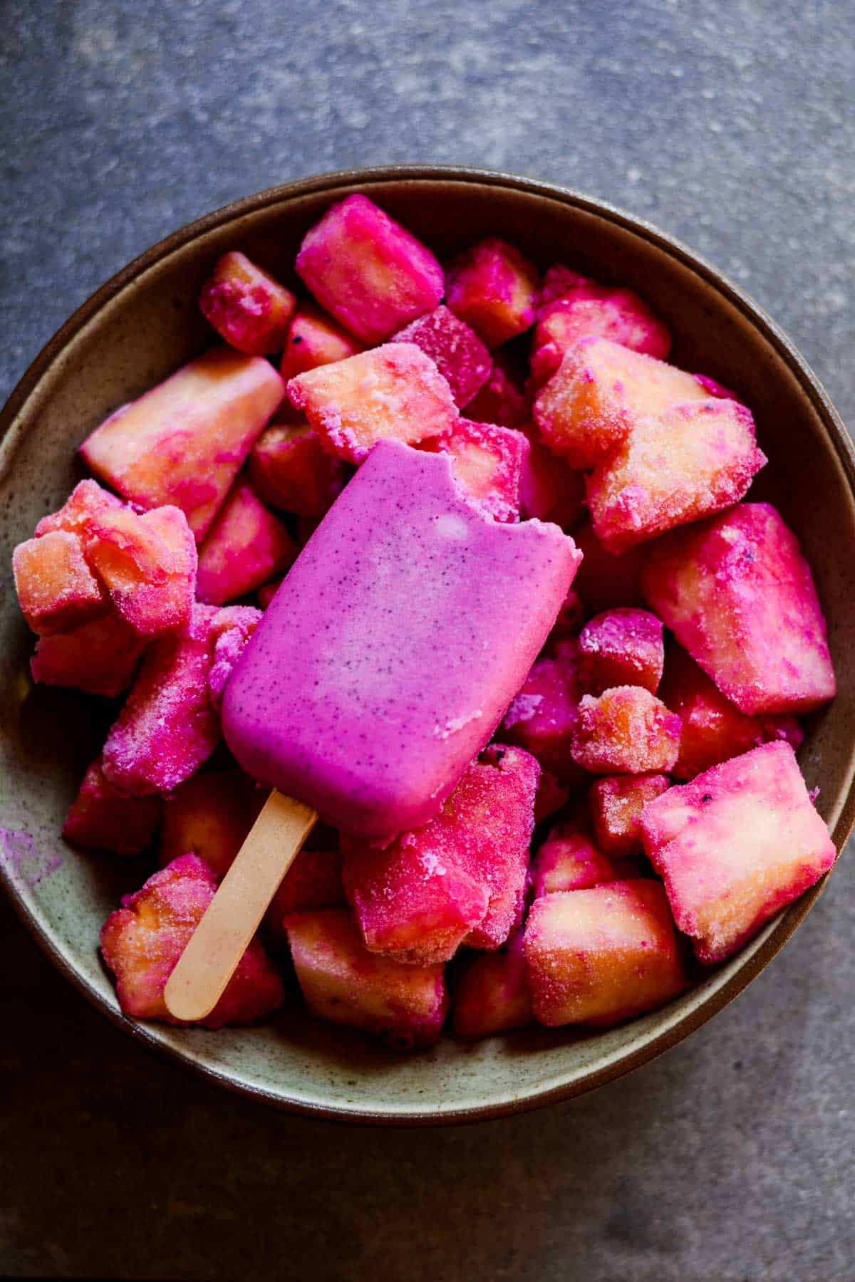 Bite out of bright purple dragon fruit popsicle, sitting on in a bowl of frozen fruit.