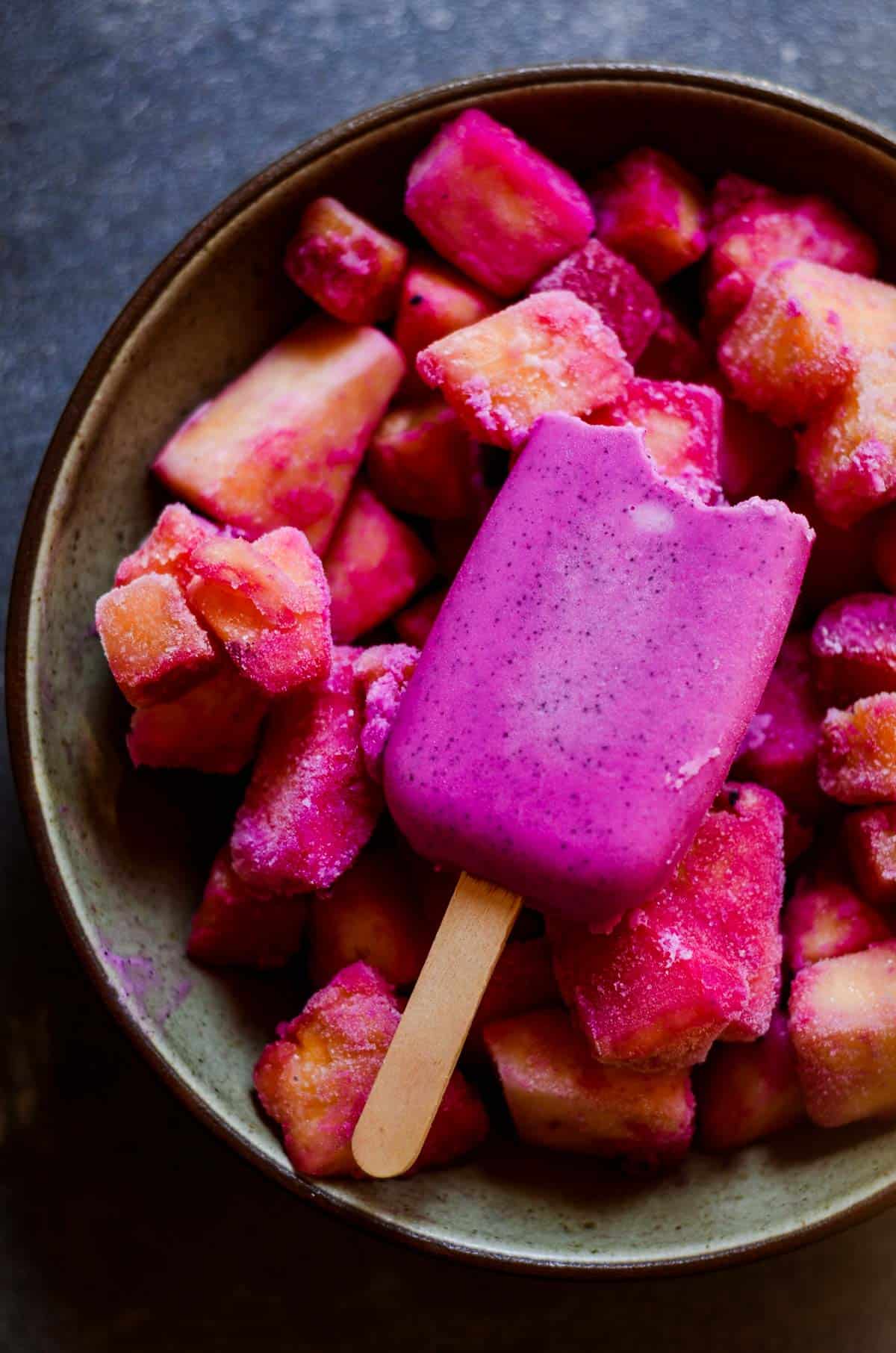 A bite from a bright purple popsicle sitting on on frozen fruit in a bowl.