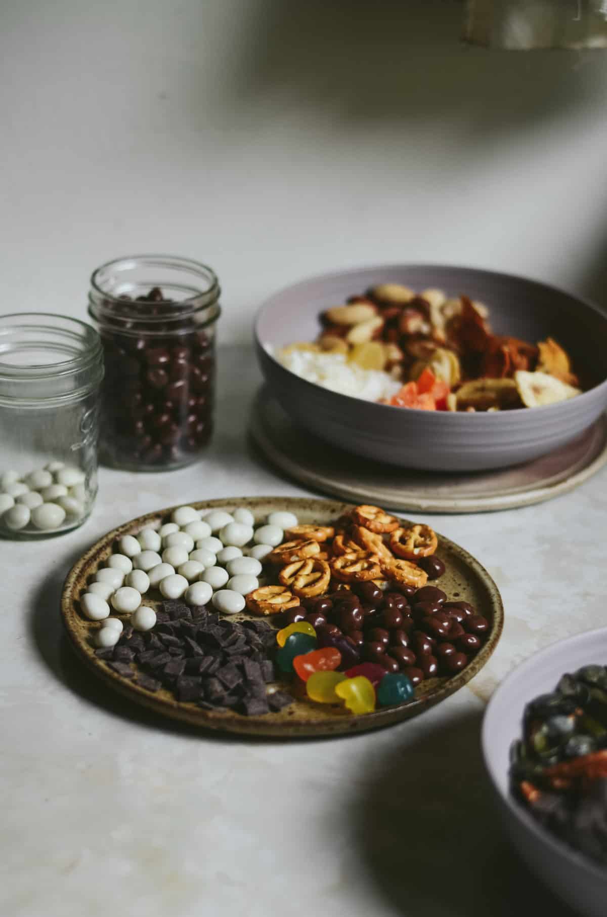 Three bowls and two jars with chocolate covered raisins, yogurt covered raisins, pretzels, candy for trail mix