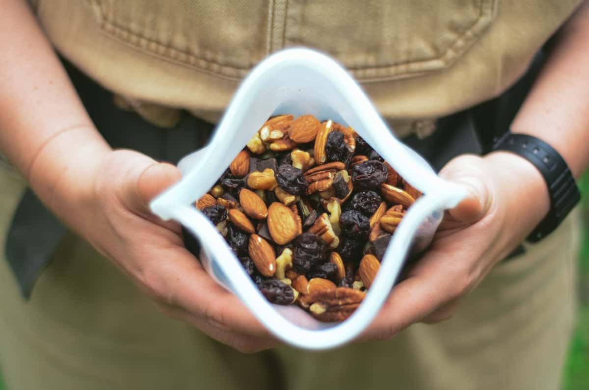 Hiker with backpack holding a bag of trail mix open with almonds, chocolate, pecans, pumpkin seeds.