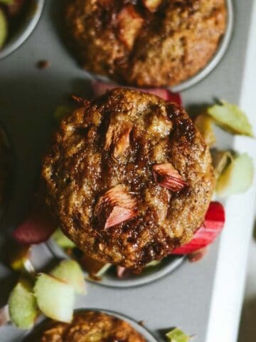 Brown muffin with pink rhubarb and brown sugar on top, sitting in a muffin tray.