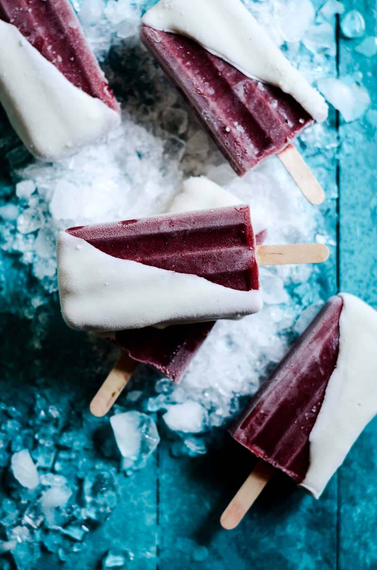 Cherry popsicles that are dark red dipped in chocolate laying on a bright blue counter with ice on it.