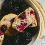 Pale brown muffins in a bowl with red blackberries.