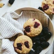 Pale brown muffins with two dark blackberries on the top, sitting in a serving bowl.