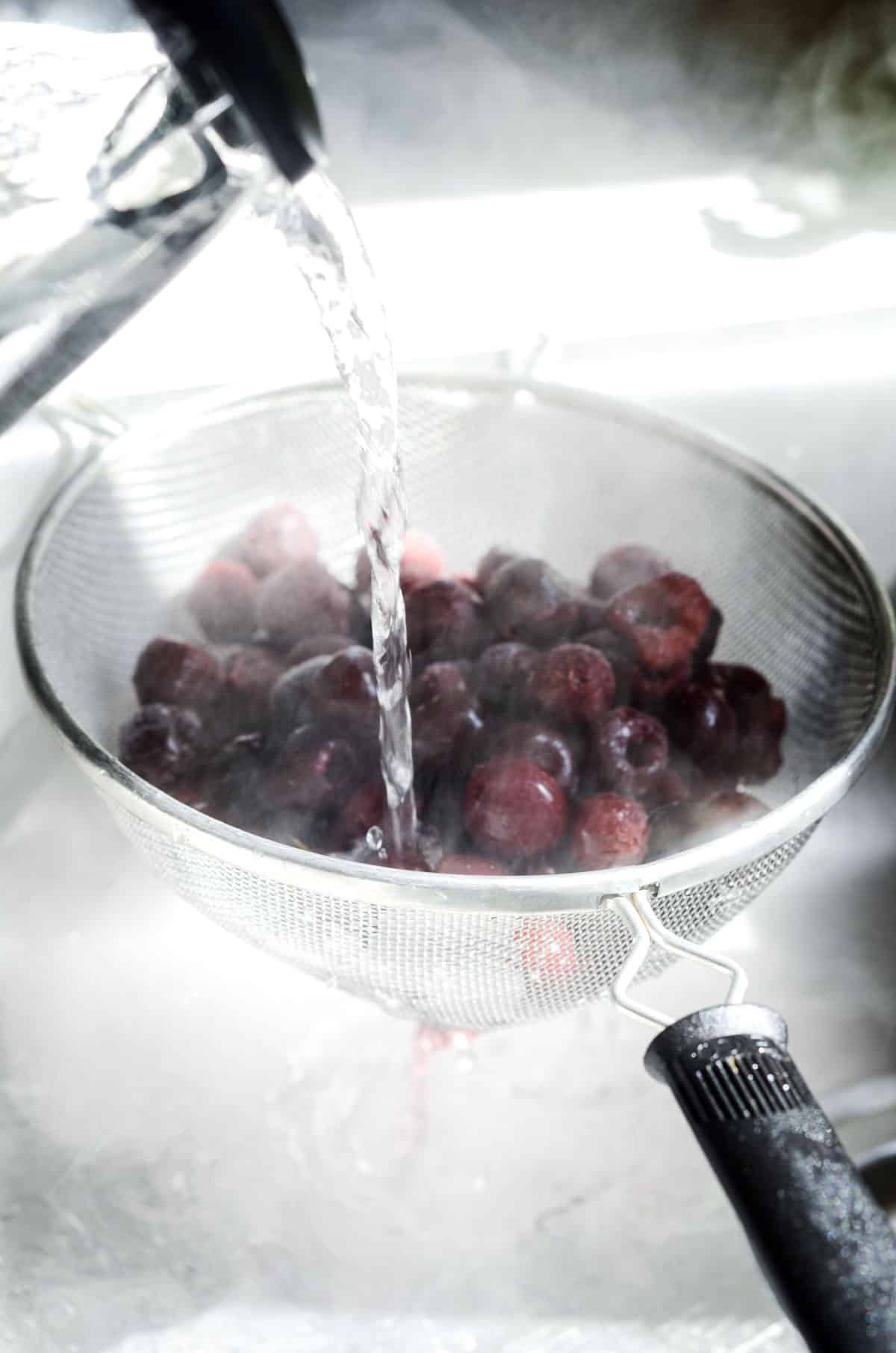 Frozen cherries in a colander with a kettle of hot water being pour onto them.