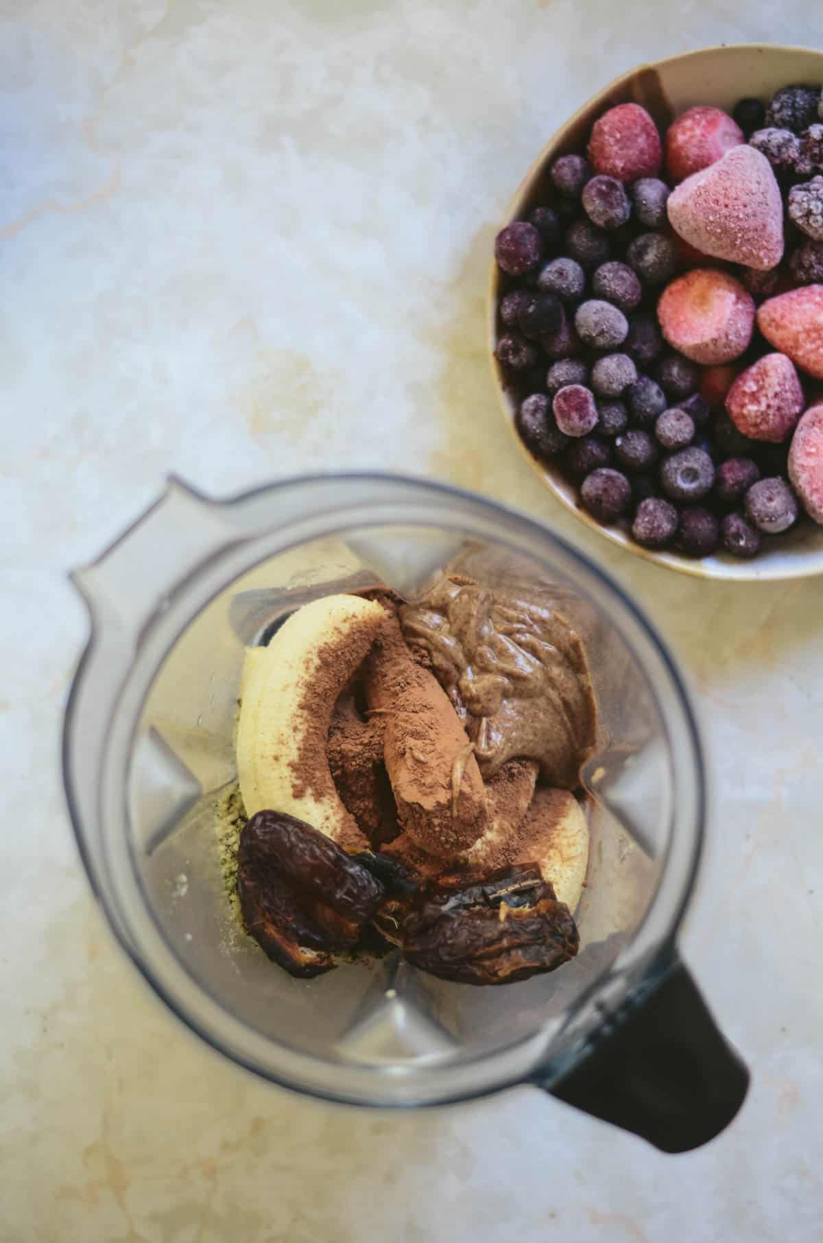 Almond butter, cocoa powder, banana, hemp hearts, dates, in the blender container with the frozen berries in a pottery bowl beside.