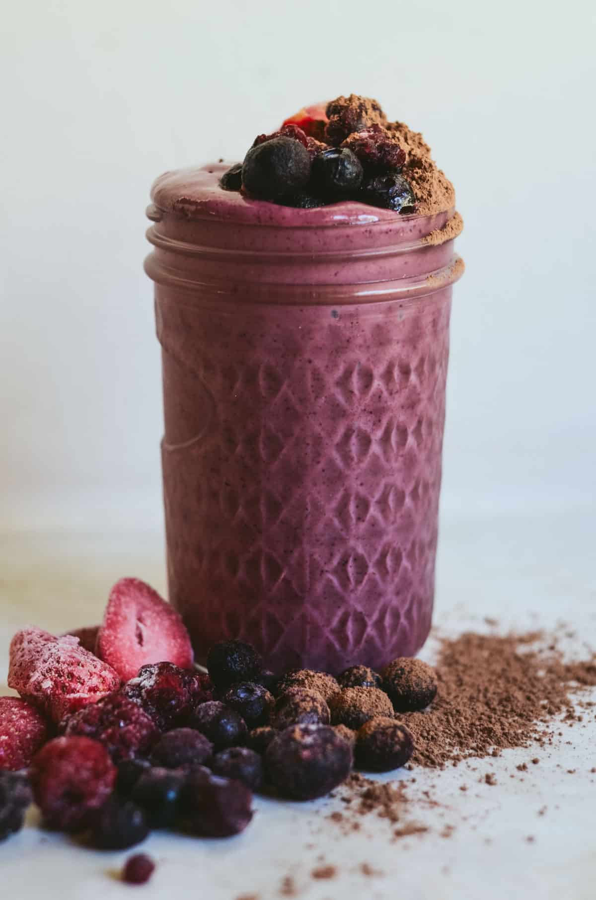 Dark purple smoothie in a glass jar with blueberries and strawberries and cocoa powder on top and on the counter.