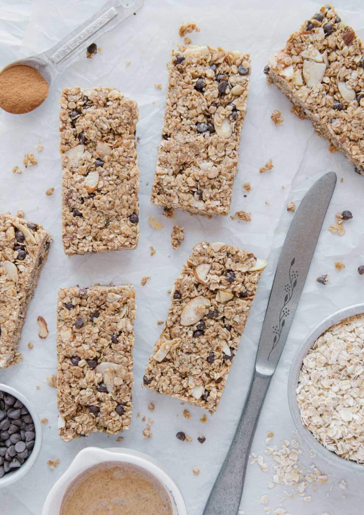 No bake granola bars sitting on the counter with almond butter and chocolate chips and oats all around it.