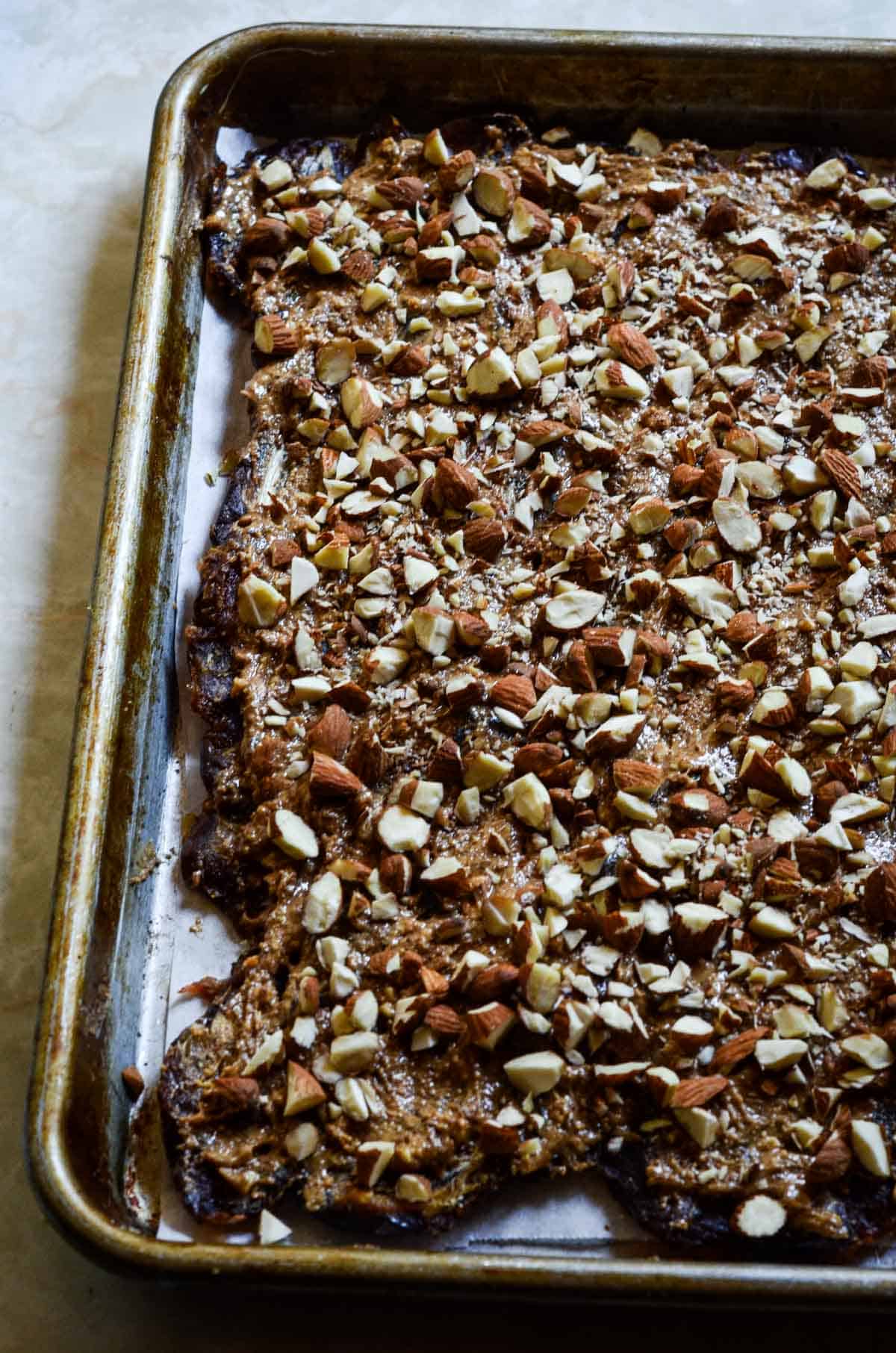 Almond butter and chopped nuts covering flattened dates on a small baking tray.