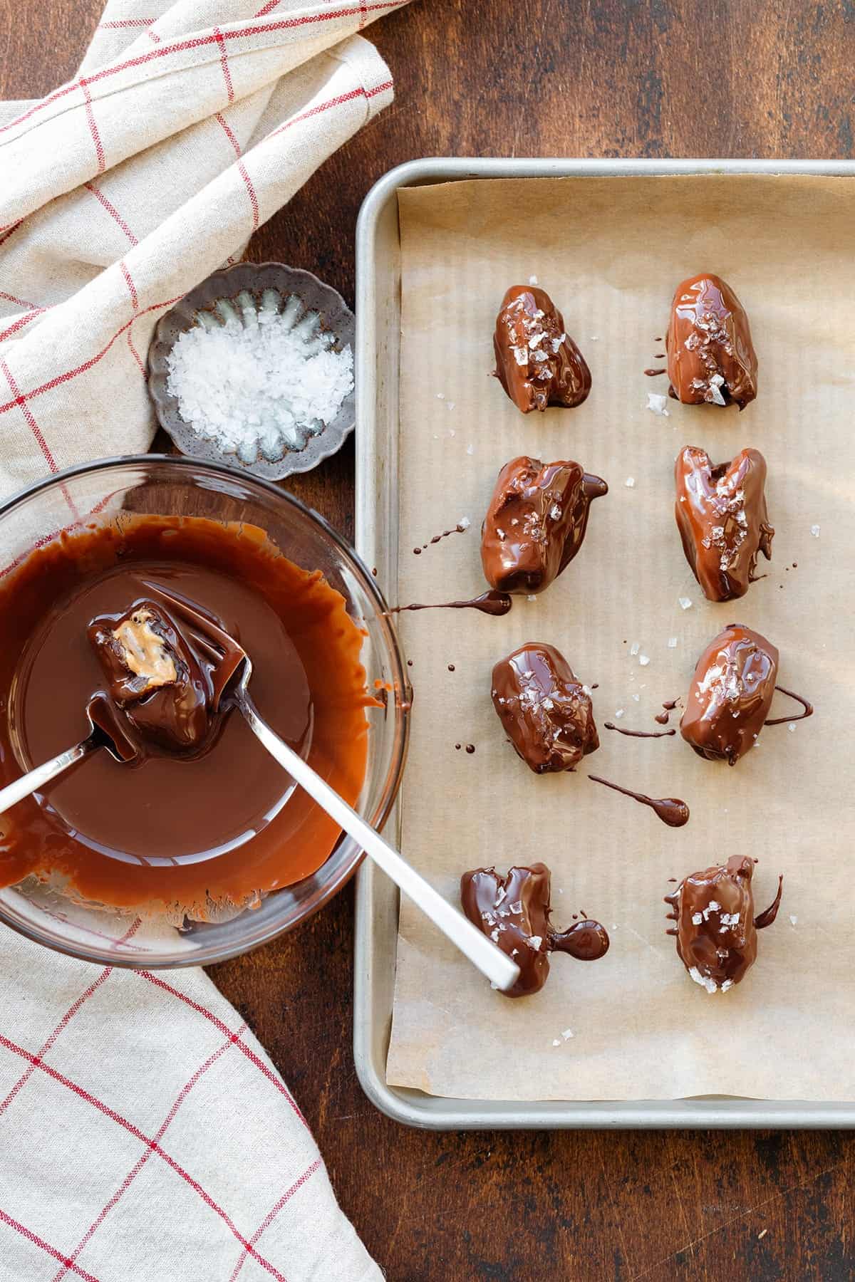 A baking pan with parchment paper on top, dates that have chocolate on them and sea salt on pan, melted chocolate and sea salt beside the pan.