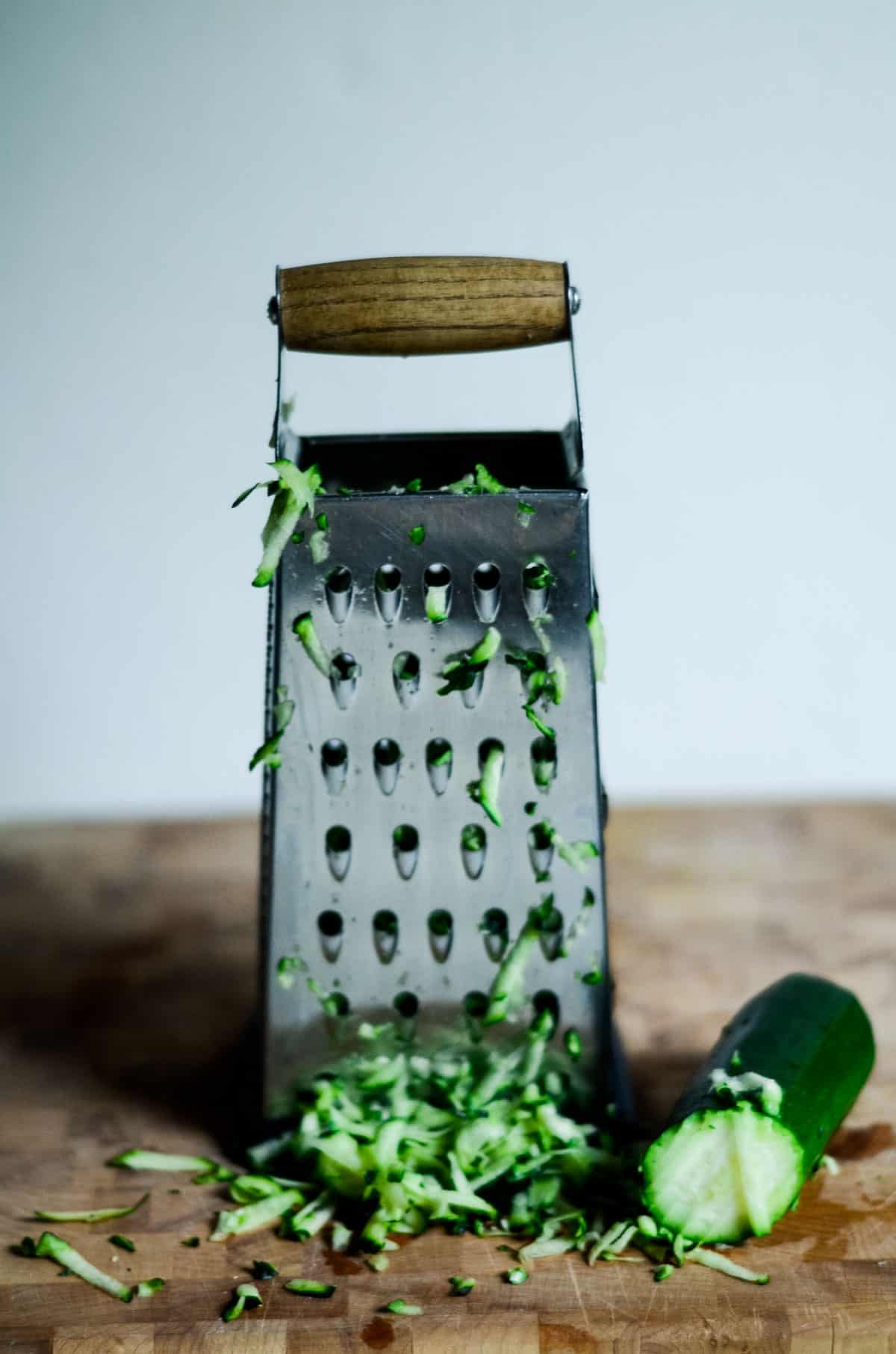 Grating zucchini on a box grater with a wooden handle, half a green and white zucchini sitting on cutting board. 