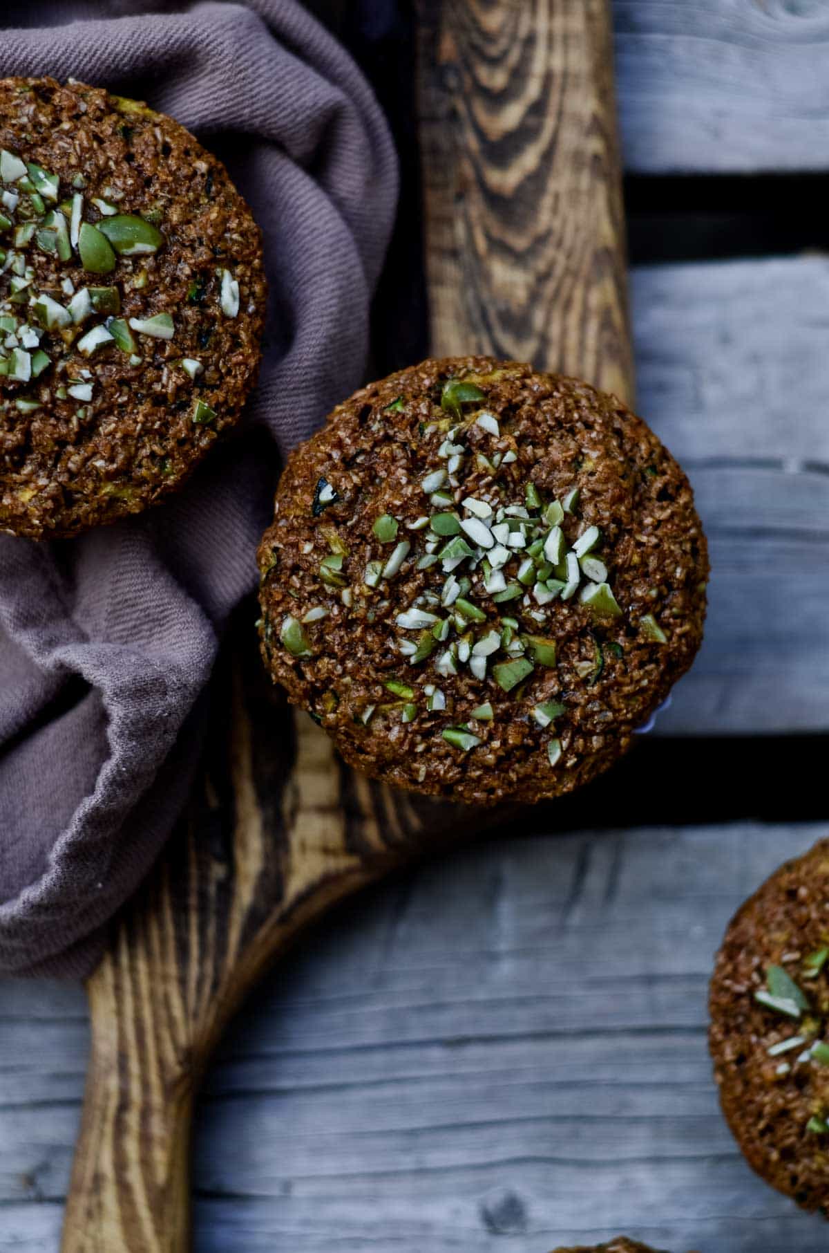 Bran muffin with green pumpkin seeds on top, on a wooden platter with 2 other muffins close by.