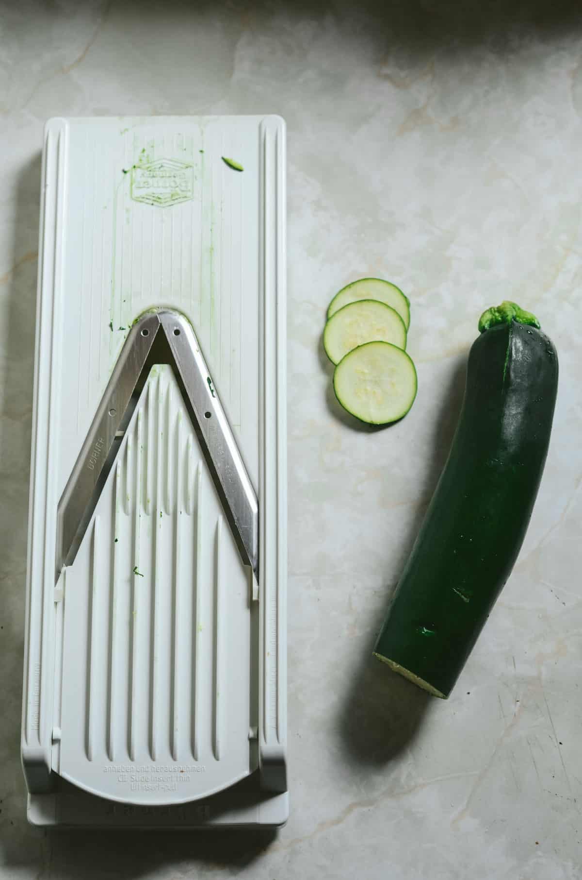 Manoline slicer with a couple of zucchini rounds beside it and half of a fresh zucchini.