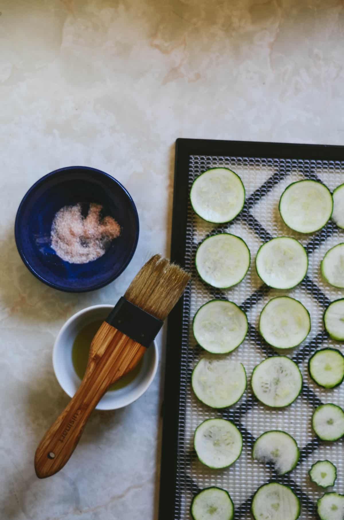 Raw zucchini rounds on dehydrator tray with a little bowl of salt and a little bowl of oil and a pastry brush beside.