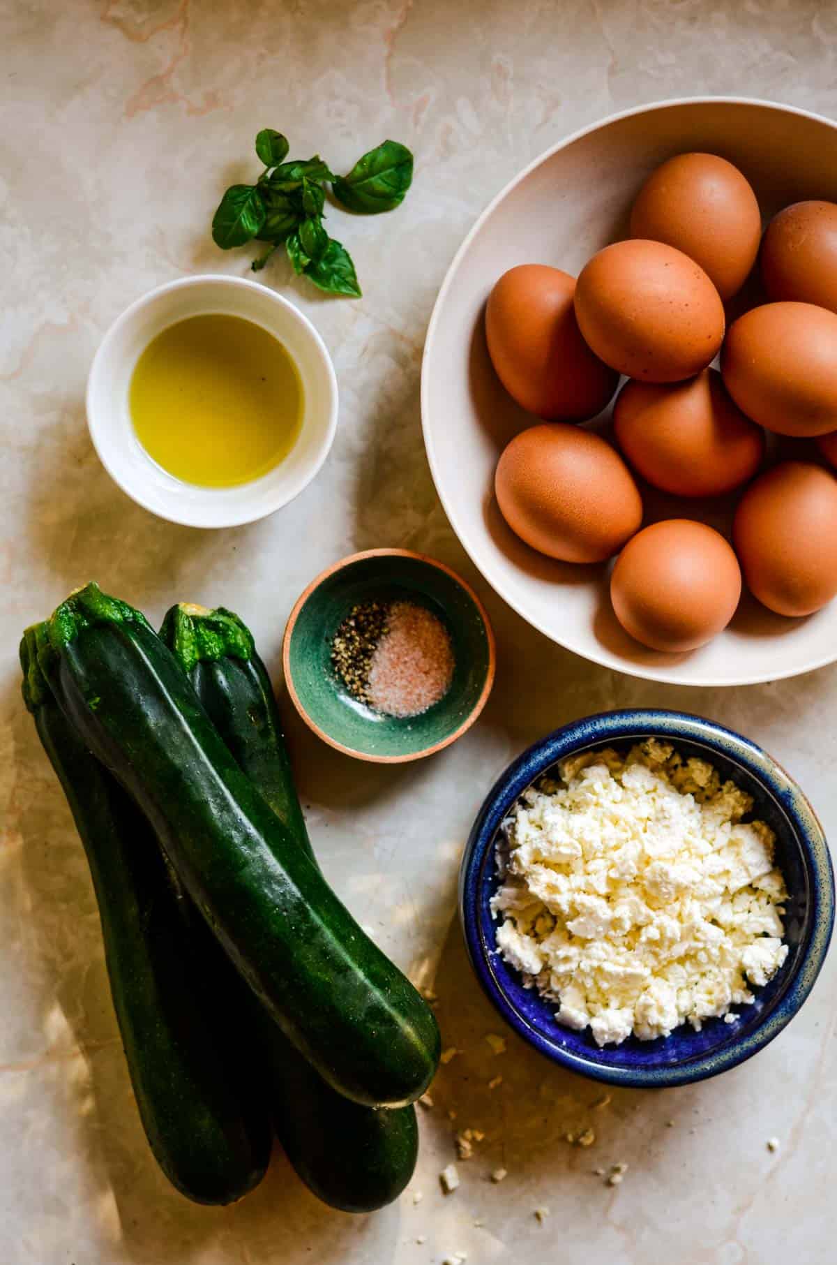 Three fresh zucchini, sea salt, oil, fresh basil, eggs, and feta in bowls measured for egg bake.