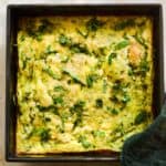 Flay lay of yellow eggs baked with green flecks of zucchini and fresh basil, and pot holder on side of pan.