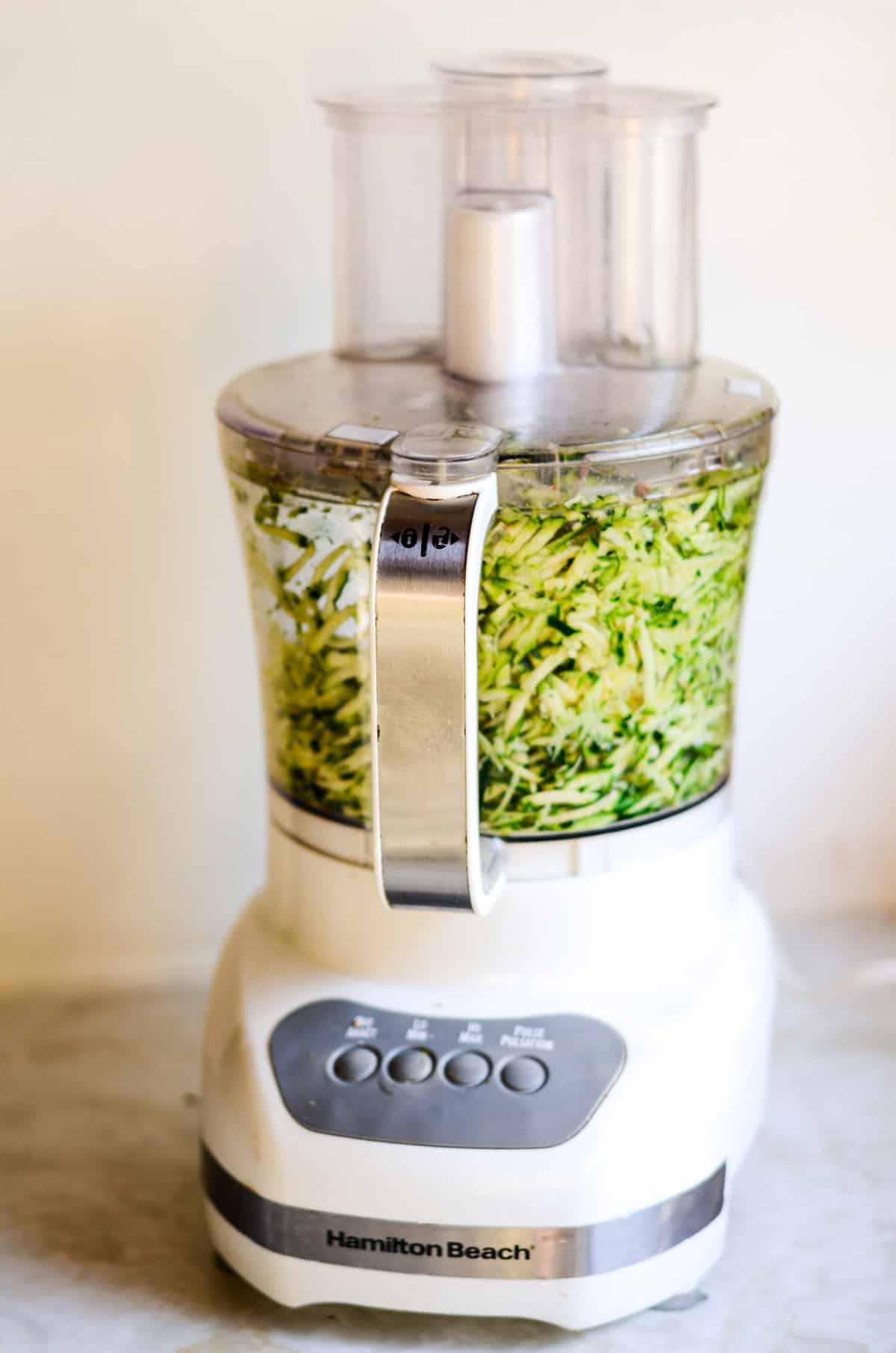 A white food processor with shredded zucchini inside.