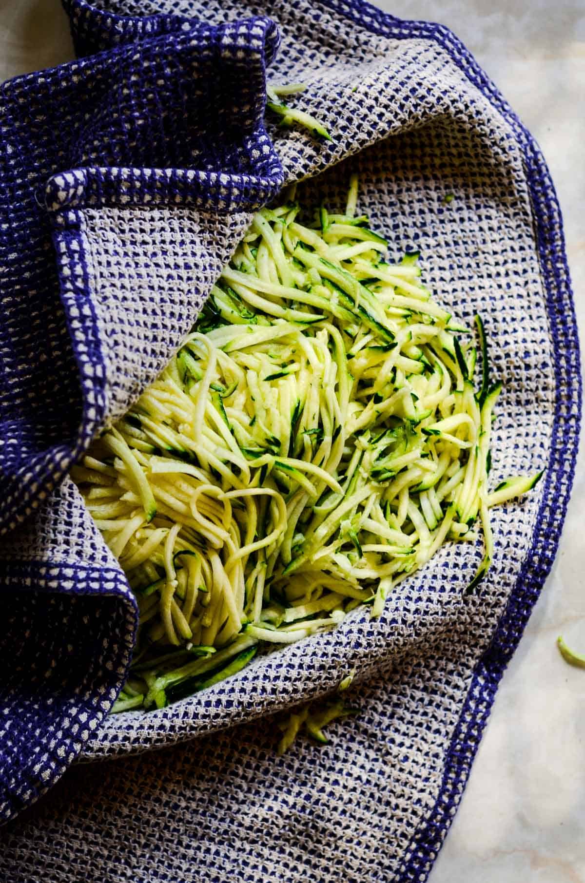 Shredded zucchini in a blue tea towel being squeezed out.