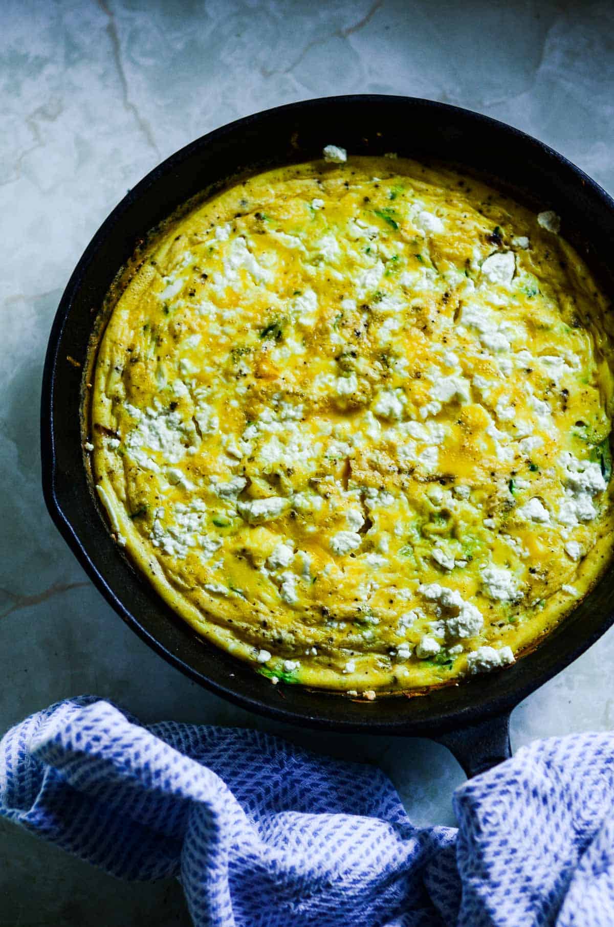 Bright yellow eggs baked in a cast iron pan with green flecks of zucchini in the eggs, and white flecks of feta cheese. 