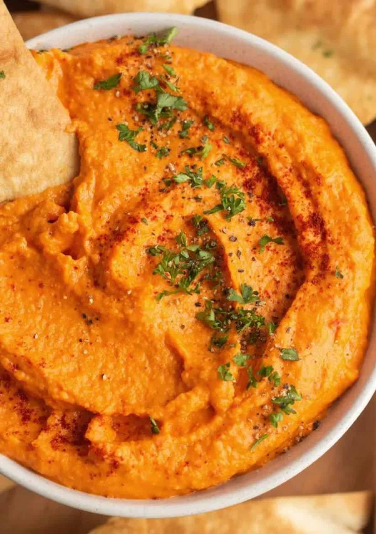 Orange colored dip with green herbs on top and a chip dipped in.