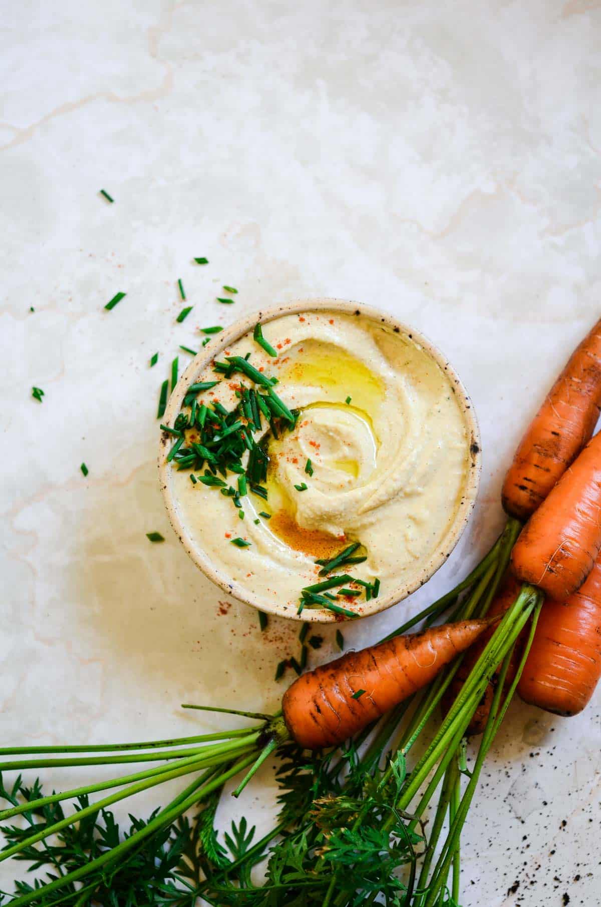 A bowl of light brown hummus with chives and oil on top, and fresh carrots with greens attached beside.