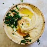 Light brown hummus in a bowl with chive and oil on top.