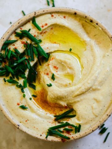 Light brown hummus in a bowl with chive and oil on top.