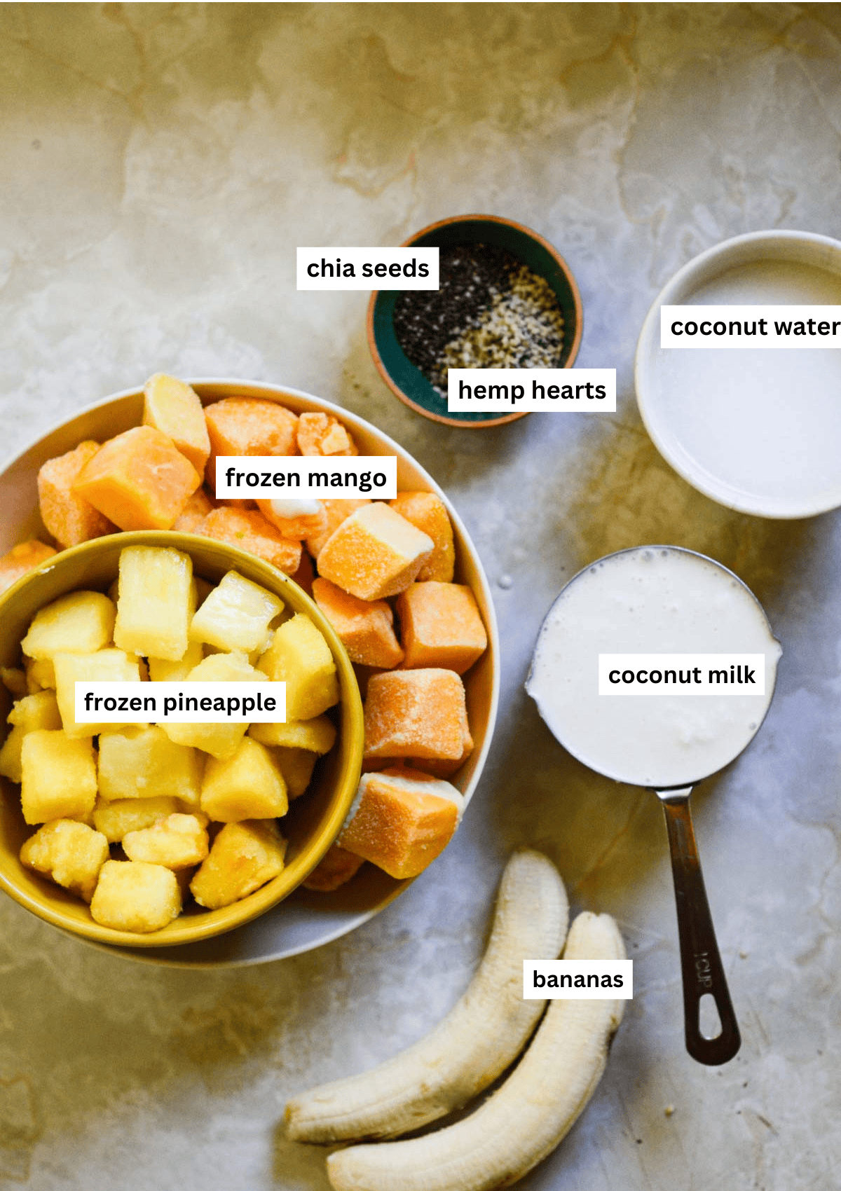 Pineapple, mango, bananas, coconut milk, coconut water, hemp hearts, and chia seeds, all in bowls.