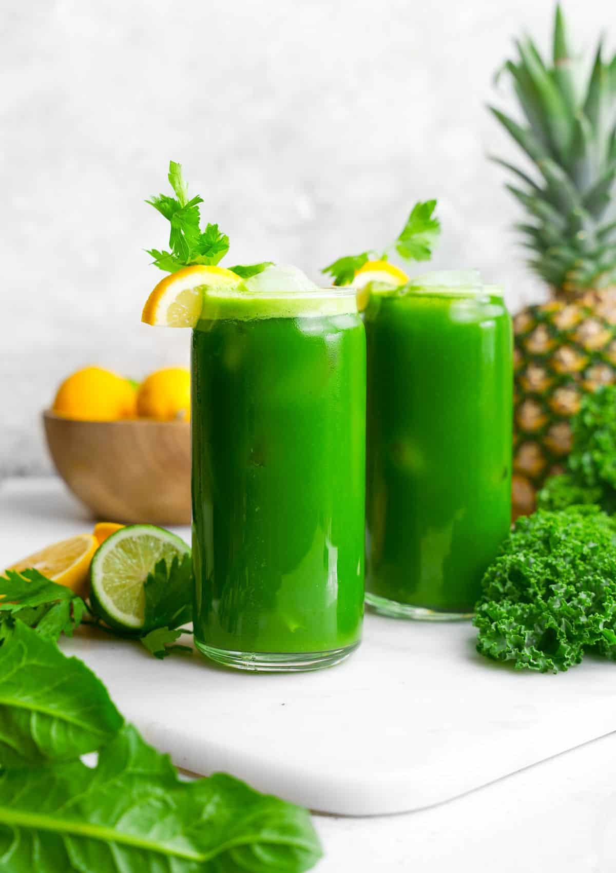 Bright green juice in two glasses with pineapple behind it.