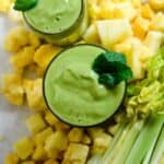 2 cups of light green smoothie in a glass cup with frozen pineapple and fresh celery stalks beside it.