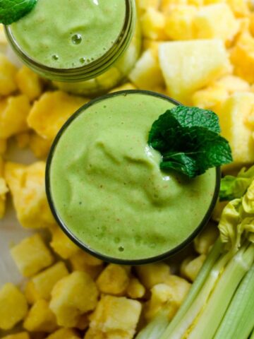 2 cups of light green smoothie in a glass cup with frozen pineapple and fresh celery stalks beside it.