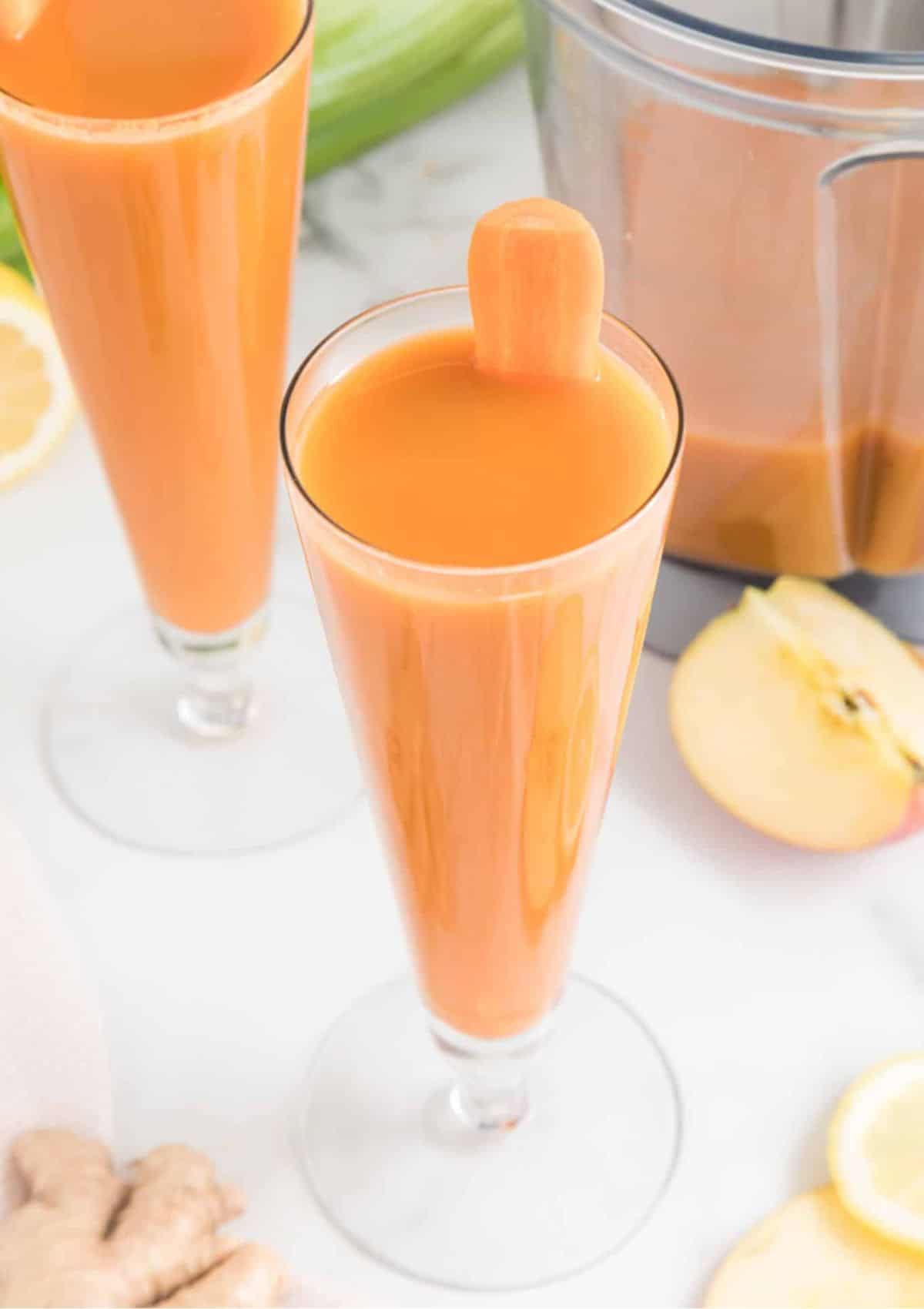 Carrot juice in two tall glasses with a carrot coming out of it.