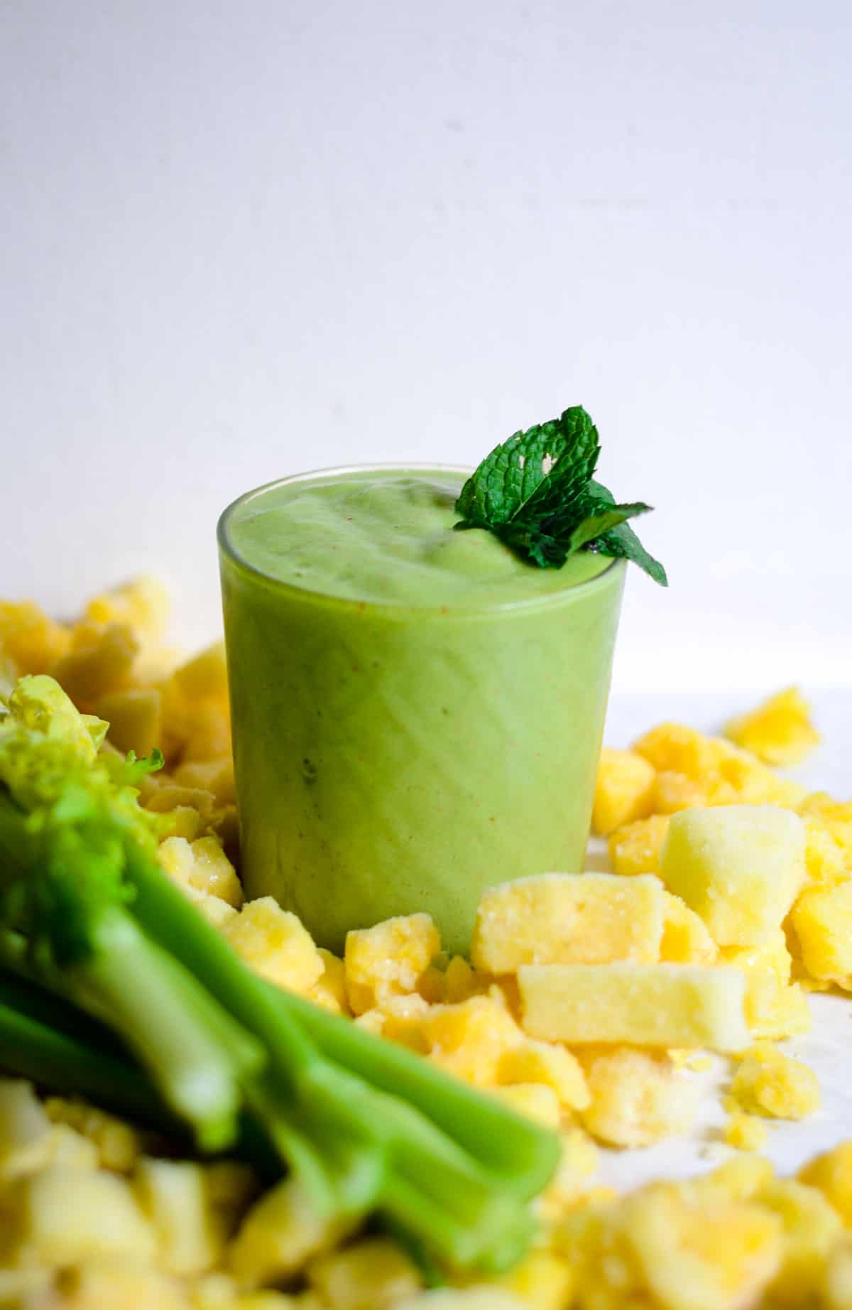 Green smoothie in a glass cup, with mint coming out of it, with frozen pineapple and fresh celery stalks beside it.