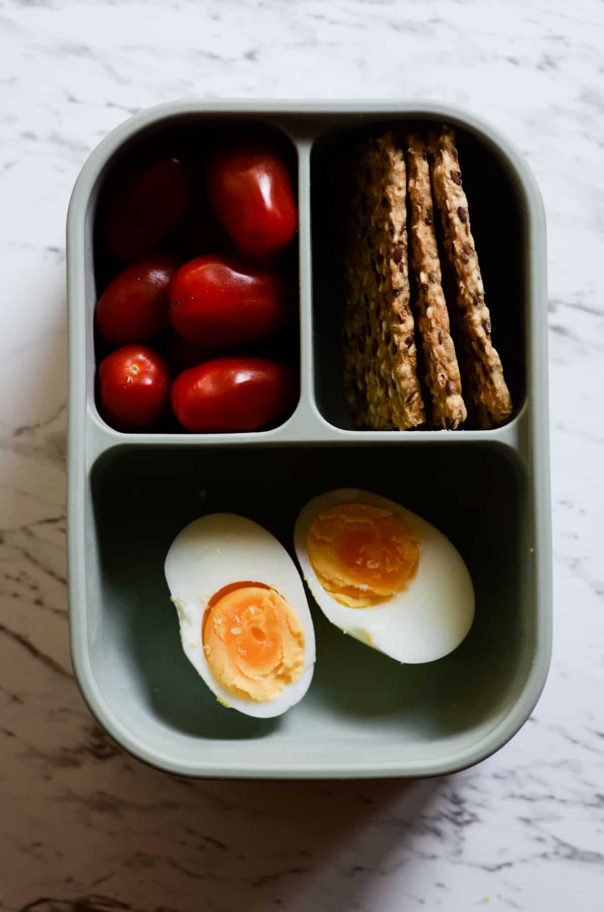 In a bento box style container, cherry tomatoes, seed crackers, and boiled eggs. 