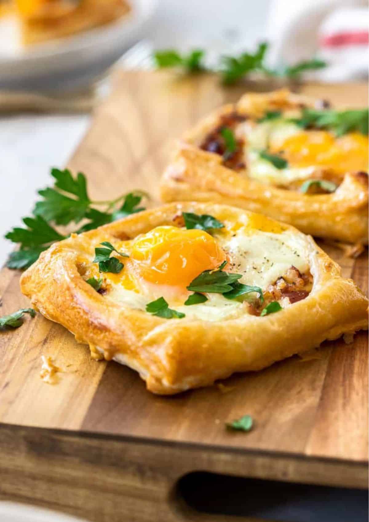 Square puff pastry with egg and bacon inside of it sitting on a cutting board with parsley. 