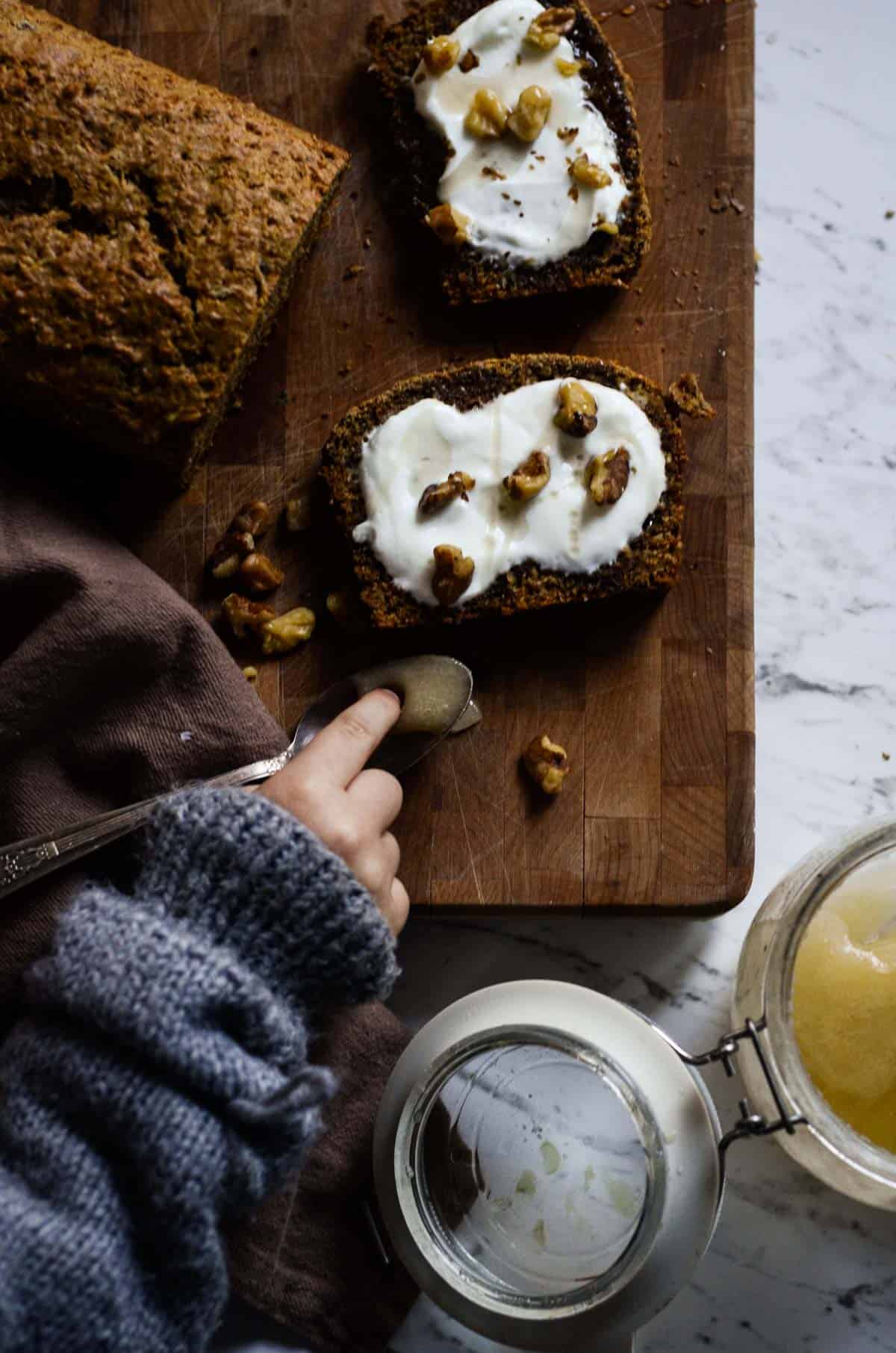 banana bread slices cut on a wood board with yogurt, nuts, and honey on top, a hand reaching the honey. 