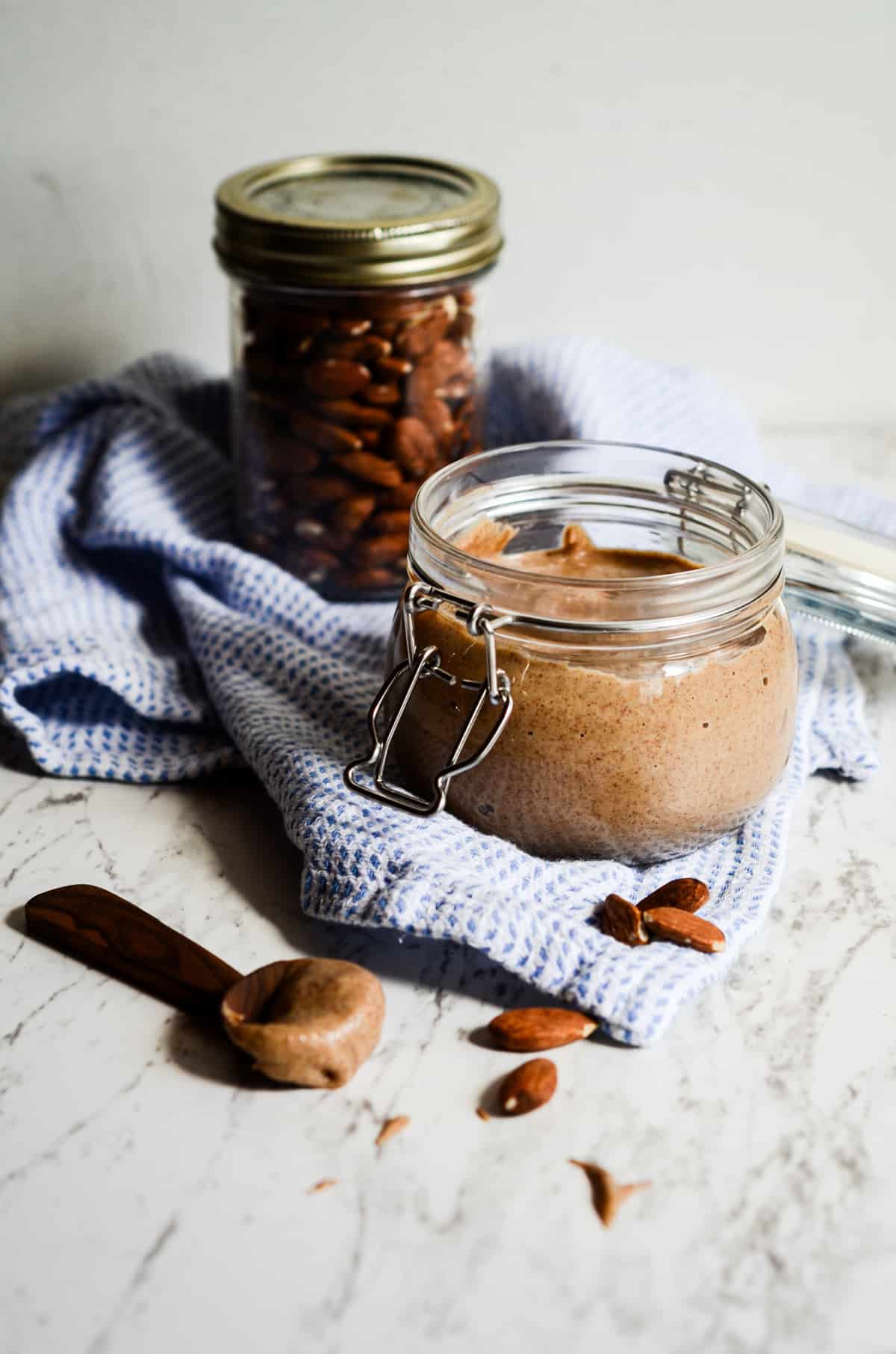 Homemade almond butter in a jar with roasted almonds beside the jar on the counter. 