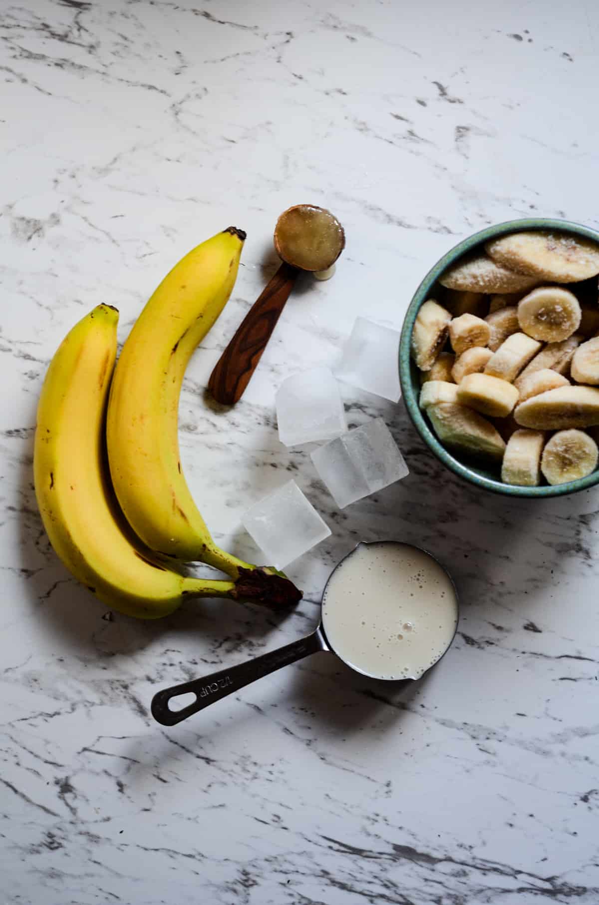 2 bananas, 1 wooden spoon of honey, a measuring cup with milk, a couple ice cubes, and a bowl or frozen banans all on the counter.