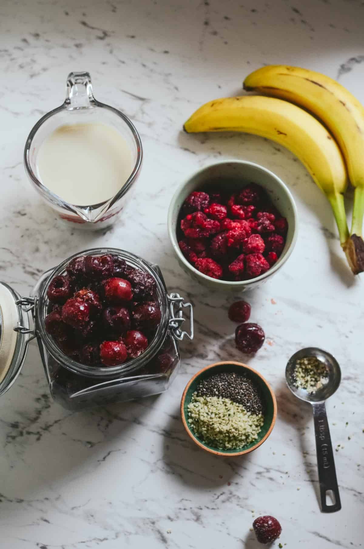 Ingredients for raspberry cherry smoothie in bowls: sweet dark cherries, frozen raspberries, almond milk, chia seeds, hemp hearts, and bananas.