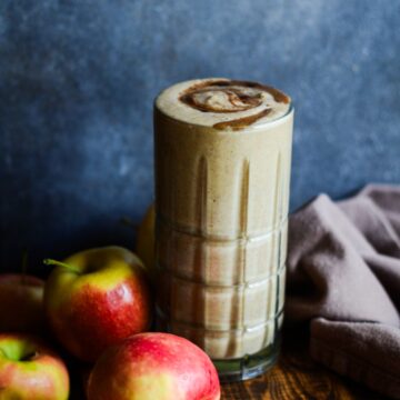 Light brown apple smoothie in tall glass with yellow and red apples beside it.