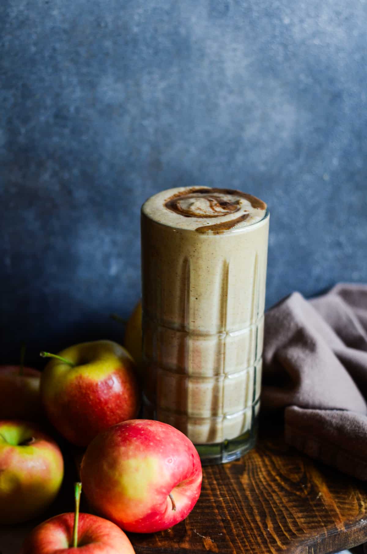 Light brown apple smoothie in a tall glass cup with an almond butter drizzle with apples beside it.